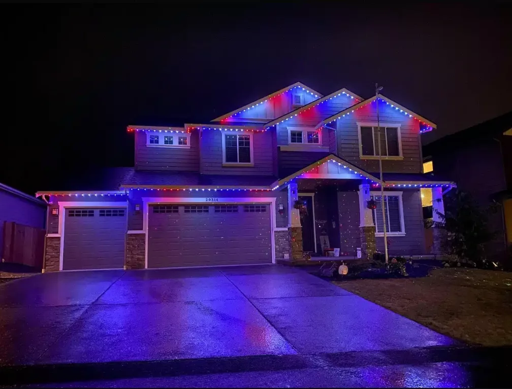 Red, white, and blue permanent holiday lighting on the outside of a two story house with a driveway 